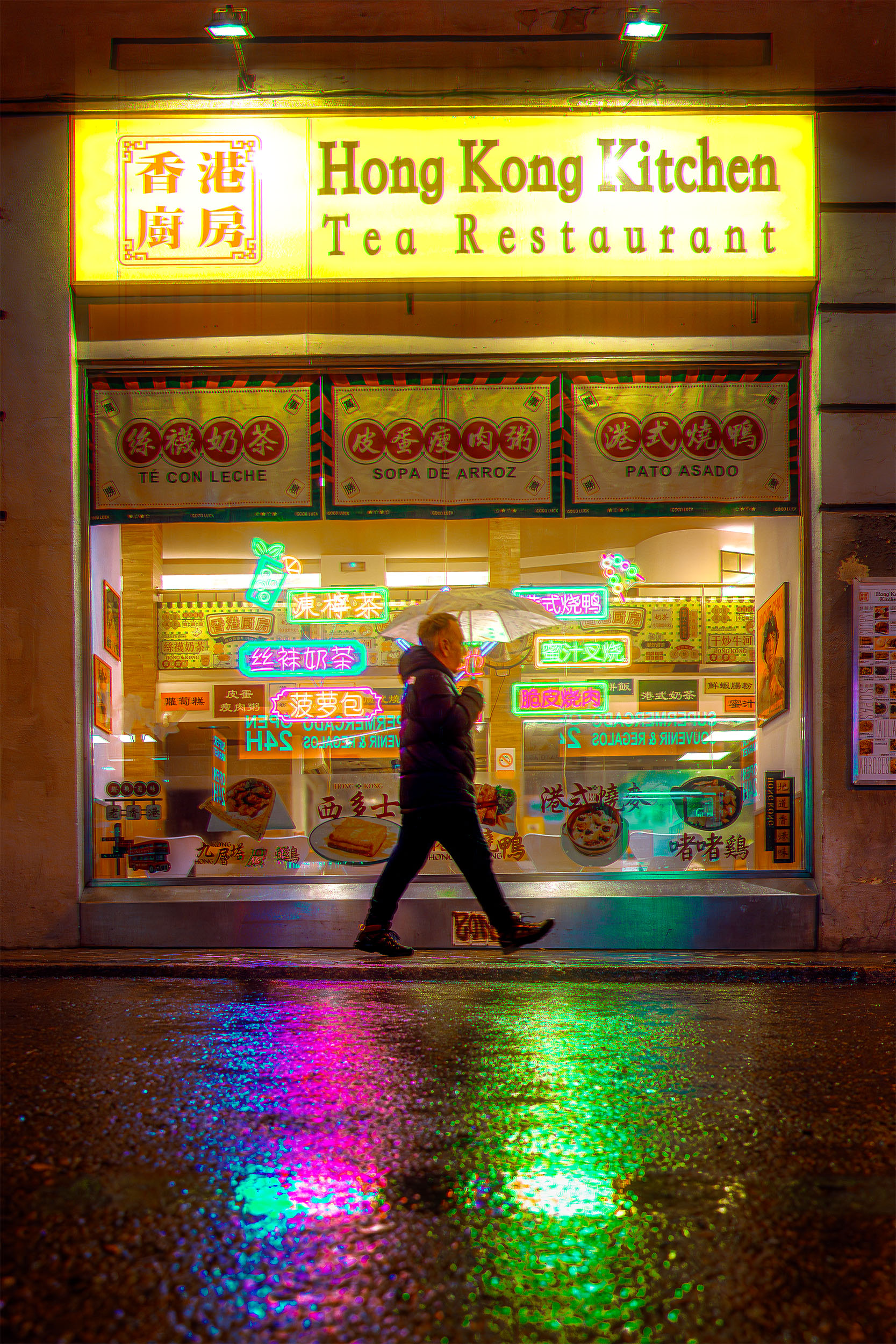 Pedestrian with umbrella passing Hong Kong Kitchen restaurant at night in Madrid, colorful neon signs reflecting on wet pavement in purple and green