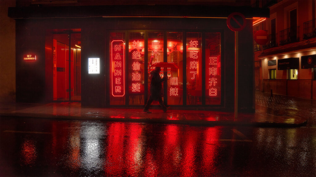 Lone figure walking past Hammer restaurant at night in Madrid, vertical red neon signs with Chinese characters reflecting on rain-soaked street