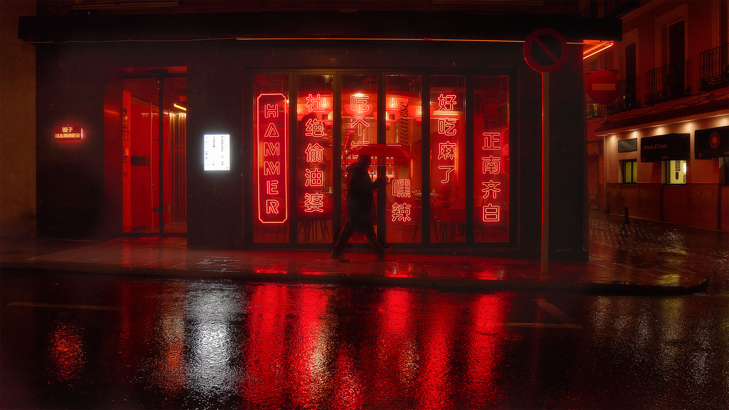 Lone figure walking past Hammer restaurant at night in Madrid, vertical red neon signs with Chinese characters reflecting on rain-soaked street
