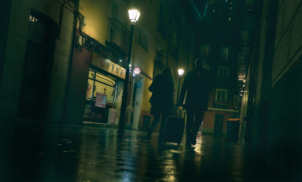 Two silhouettes with luggage walking through a narrow rain-soaked Madrid alley at night, warm street lamps reflecting on wet cobblestones