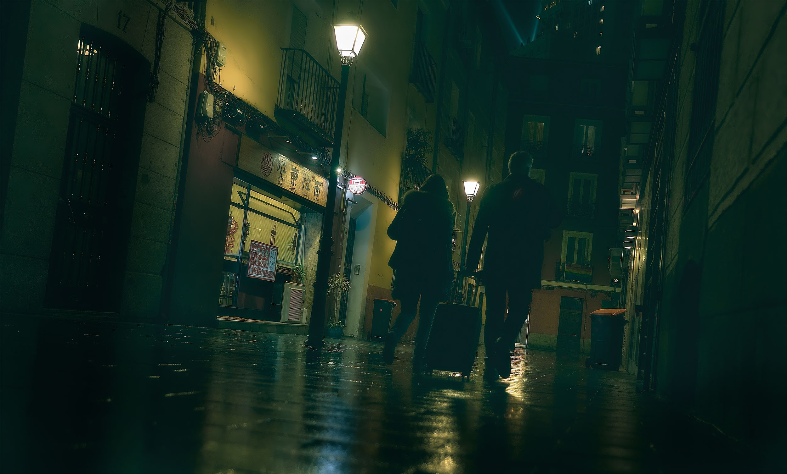 Two silhouettes with luggage walking through a narrow rain-soaked Madrid alley at night, warm street lamps reflecting on wet cobblestones