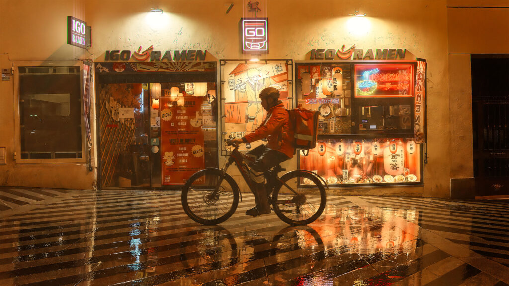 Delivery cyclist passing Igo Ramen restaurant at night in Madrid, warm orange neon light reflecting on wet street