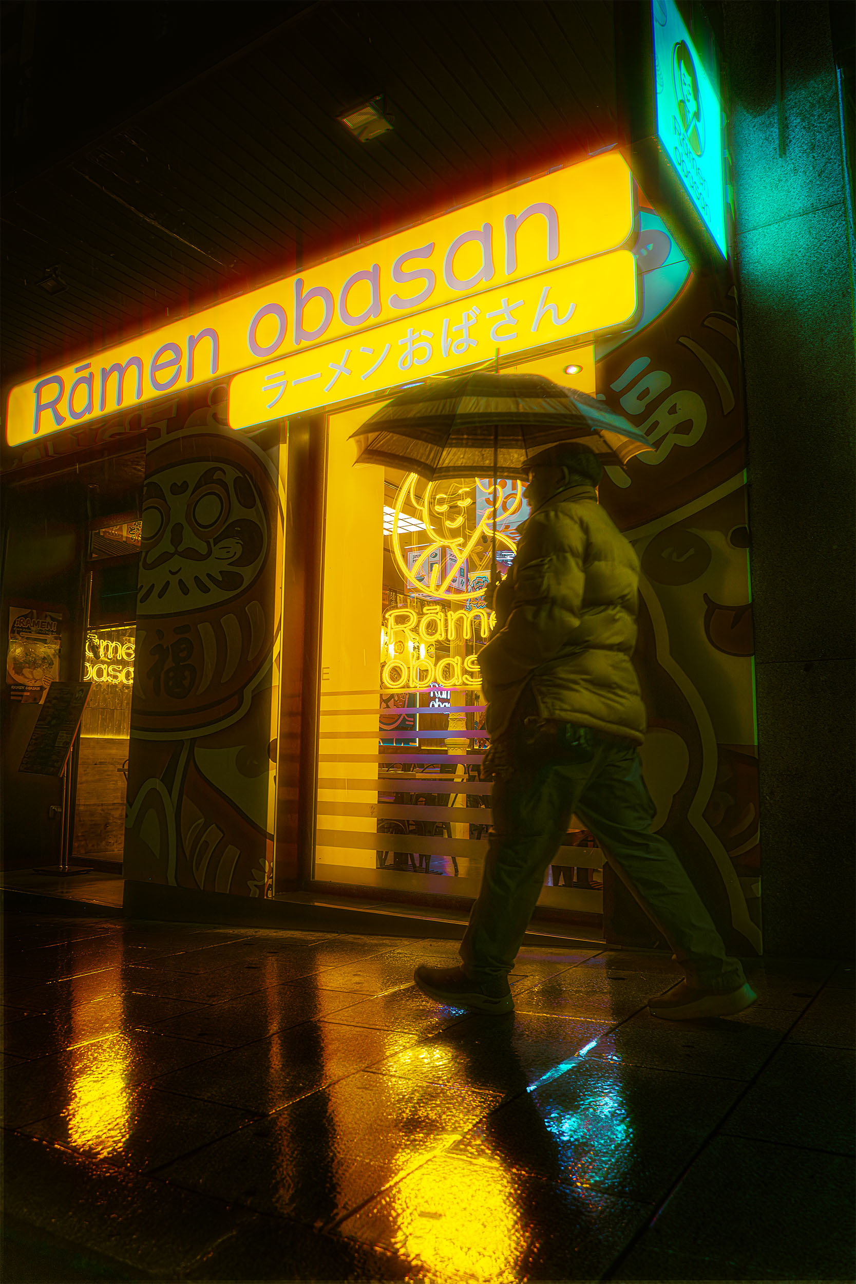 Silhouette with umbrella passing Ramen Obasan restaurant at night in Madrid, intense yellow neon sign with Japanese characters, wet pavement reflections