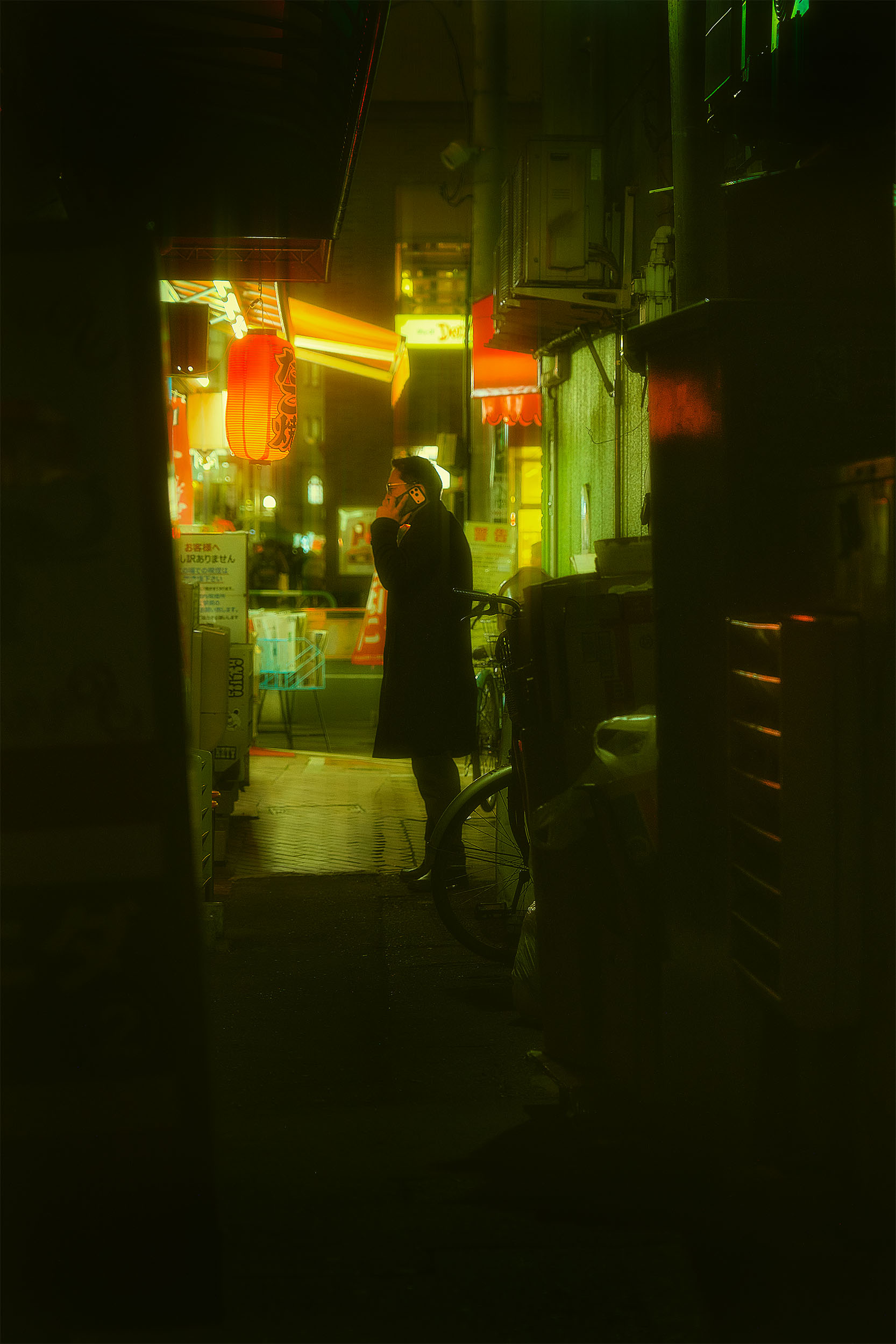 Person on a phone call standing in a Tokyo alley at night, framed by darkness, red lantern and neon signs glowing in the background, green atmospheric haze