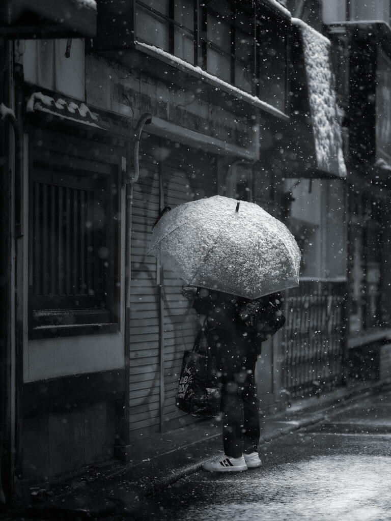 Person sheltering under a transparent umbrella during a heavy Tokyo snowstorm, black and white street photography
